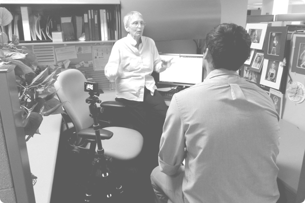 A senior woman explaining to an interviewer how she places print order online in her cubilble. A camera on a tri-pod records the interview and activity