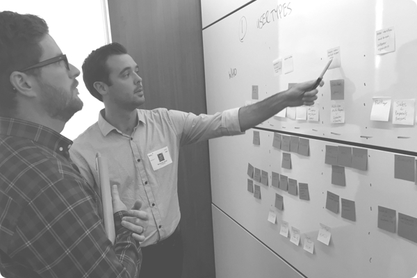 Two men point at a white board with sticky notes calling out important information