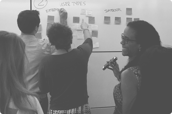 Group of employees collaborating placing post it notes onto a whiteboard. A female employee is holding up her phone smiling.