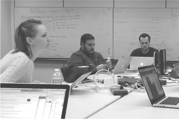 3 individuals sit at a table with laptops open, surrounded by white boards thinking pensively.