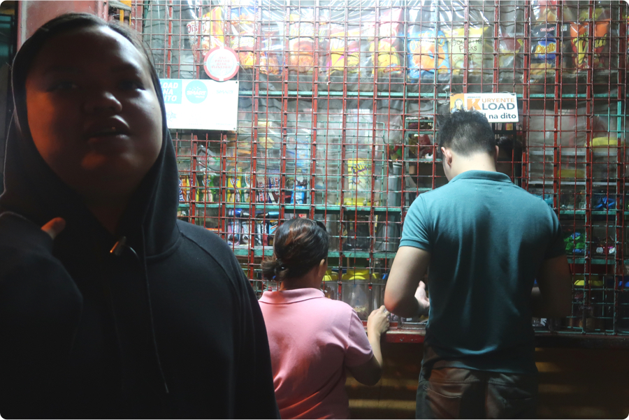 Backlit individual standing in front of a Sari Sari with bars on the store front. A man and a woman interact with the clerk at the stand.