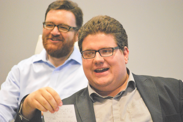 Two men smiling, one holding a piece of paper and reading aloud